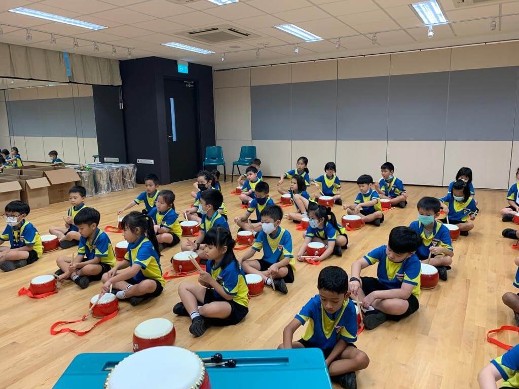 Primary school pupils sitting on a hall floor, each with a small red Chinese drum and a pair of drumsticks.