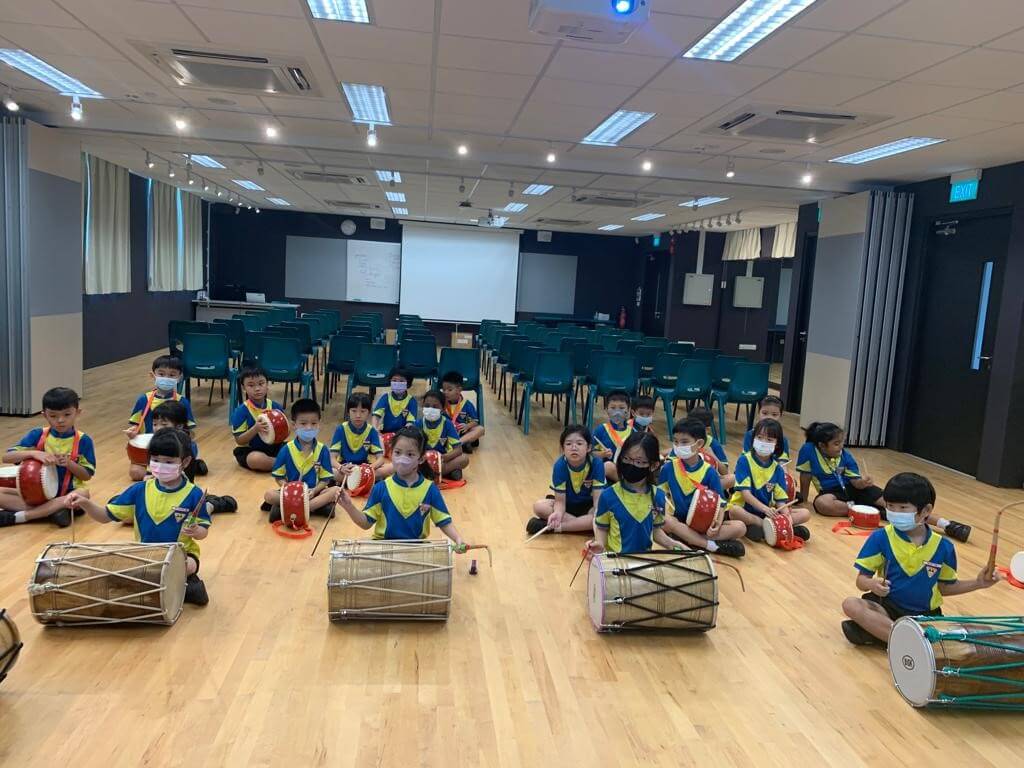 Pupils in a school hall with small red Chinese drums, and large double-headed drums laid on the floor in front of them.