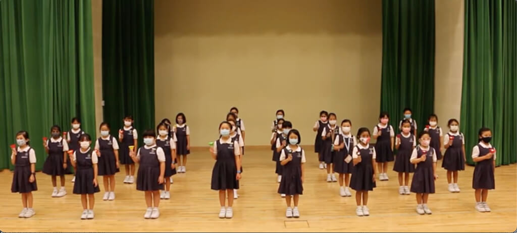 Primary school girls standing in two groups on a stage, each holding a small coloured handbell.