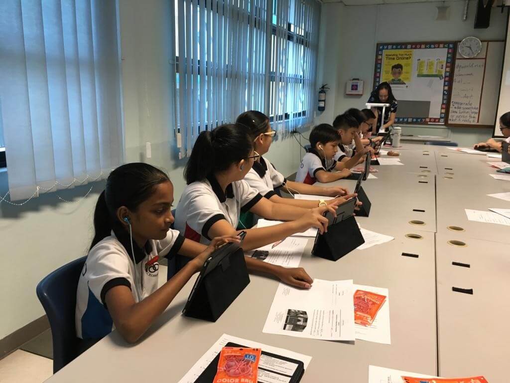 Students at a long classroom table, each working on a tablet with earphones in.