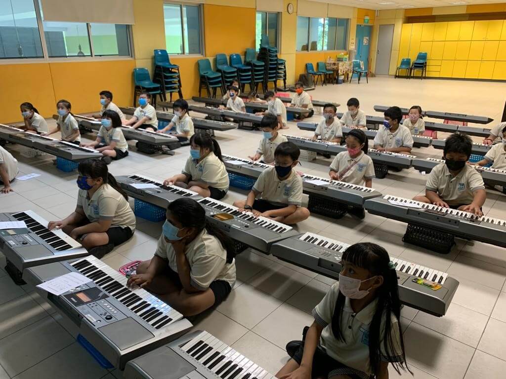 Primary school pupils sitting on the floor, each at an electronic keyboard set on a low stand.