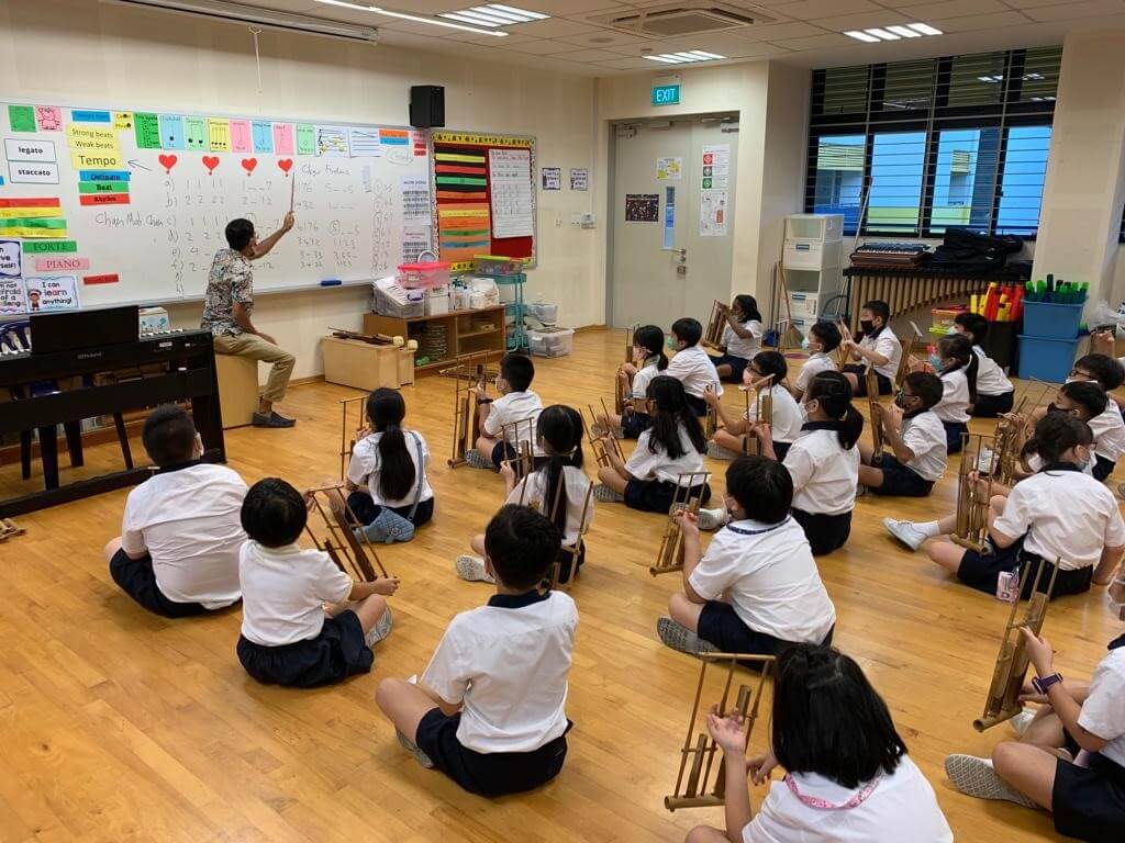 Pupils sitting on a classroom floor, each holding an angklung, while a teacher at the whiteboard points to the beat pattern.