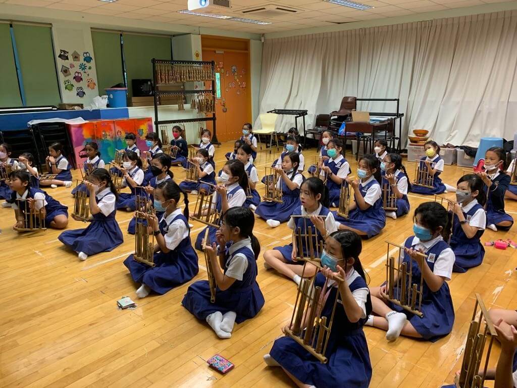 Primary school pupils sitting on a hall floor, each holding an angklung up in front of them.