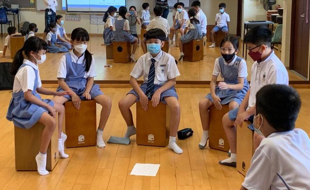 Pupils sitting on wooden cajons in a music room, with more pupils reflected in a mirror behind them.