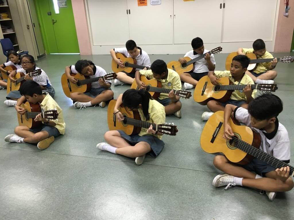 Primary school pupils sitting cross-legged on a classroom floor, each playing a classical guitar.