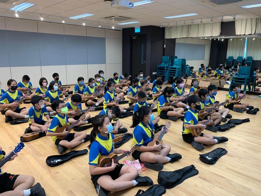 Rows of primary school pupils sitting on a hall floor, each holding a ukulele, with soft cases beside them.