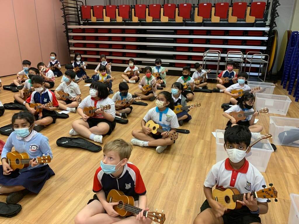 Pupils in different school uniforms sitting on a hall floor, each playing a ukulele, with tiered seating behind them.